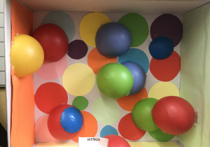 Praca przedstawia kartonowe pudełko. W środku przyklejone są kolorowe, plastikowe piłki.