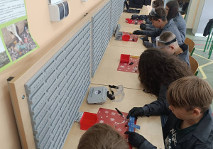 Uczestnicy warsztatów wraz z asystentami z koła „Laboratorium Przyszłości” poznają elementy zestawu, w tym płytkę PCB w kształcie satelity oraz komponenty elektroniczne.