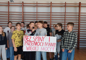 Występ klasy 6d. W tle grupa uczniów trzyma plakat o przeciwdziałaniu przemocy rówieśniczej.