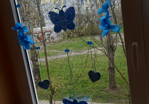 Prace uczniów przedstawiające motyle i kwiaty przyklejone do okna na szkolnym korytarzu.