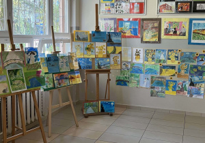 Zdjęcie wszystkich prac zamieszczonych na wystawie, na szkolnym korytarzu. Widok na sztalugi i ścianę z wyeksponowanymi pracami.