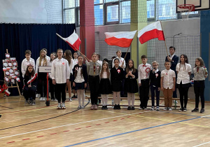 Szkolny chór, w tle uczniowie trzymający flagi Polski