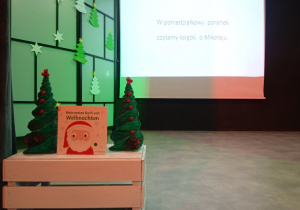 Tytułowy slajd pochodzący z prezentacji multimedialnej do przedsięwzięcia, które odbyło się 18 grudnia 2023 r. Na pierwszym planie książka o Mikołaju w języku niemieckim i świąteczne choinki.