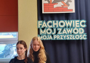 Uczennice klasy 7c stoją w głównym holu Zespołu Szkół Budowlano - Technicznych w Łodzi. W tle napis – Fachowiec, Mój Zawód, Moja Przyszłość.