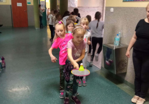 Dzieci z klas młodszych podczas zawodów sportowych na szkolnym korytarzu. Dziewczynka biegnie z paletką podczas wyścigu rzędów.