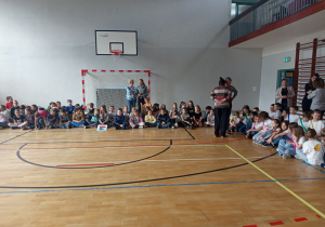 Uczniowie siedzą skrzyżnie i obserwują występy poszczególnych klas.