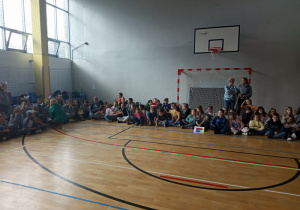 Uczniowie siedzą skrzyżnie i obserwują występy poszczególnych klas.