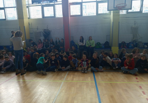 Uczniowie siedzą skrzyżnie i obserwują występy poszczególnych klas.