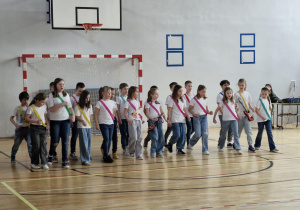 Uczniowie klasy 2c, przepasani kolorowymi szarfami, śpiewają piosenkę „Krajka”
