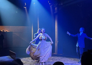 Grupa Aktorów występuje na scenie teatralnej wypełnionej ziemią.