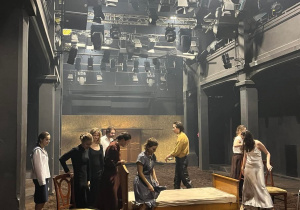 Aktorzy podczas występu w Teatrze Studyjnym, zgromadzeni wokół drewnianego łóżka.
