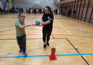 Uczestnicy zabawy mają za zadanie przenoszenie w parach piłki , która ułożona jest na dwóch laskach gimnastycznych.