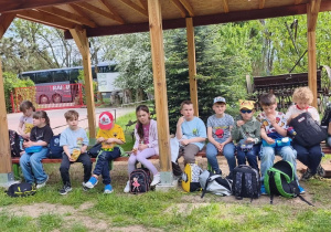 Dzieci siedzą na ławce pod altanka i jedzą prowiant, który zabrały na wycieczkę.